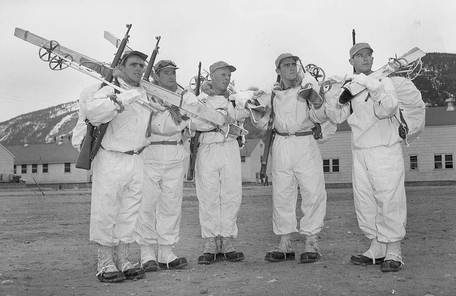 Cinco soldados de macacão branco de neve, com esquis e fuzis ao ombro, diante de alojamentos de madeira e uma montanha com neve
