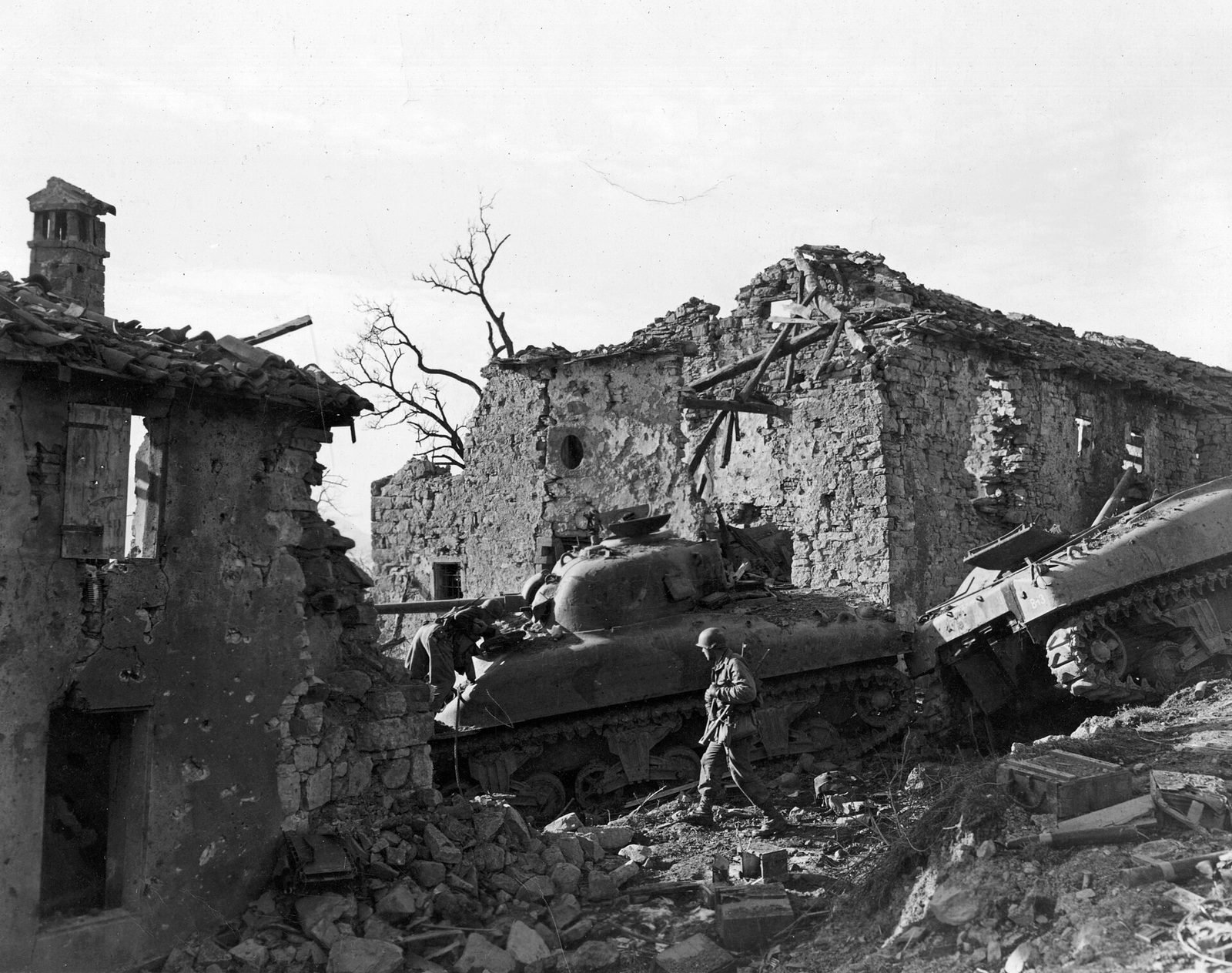 Soldado americano caminha entre casas de pedra destruídas e dois tanques Sherman avariados numa encosta