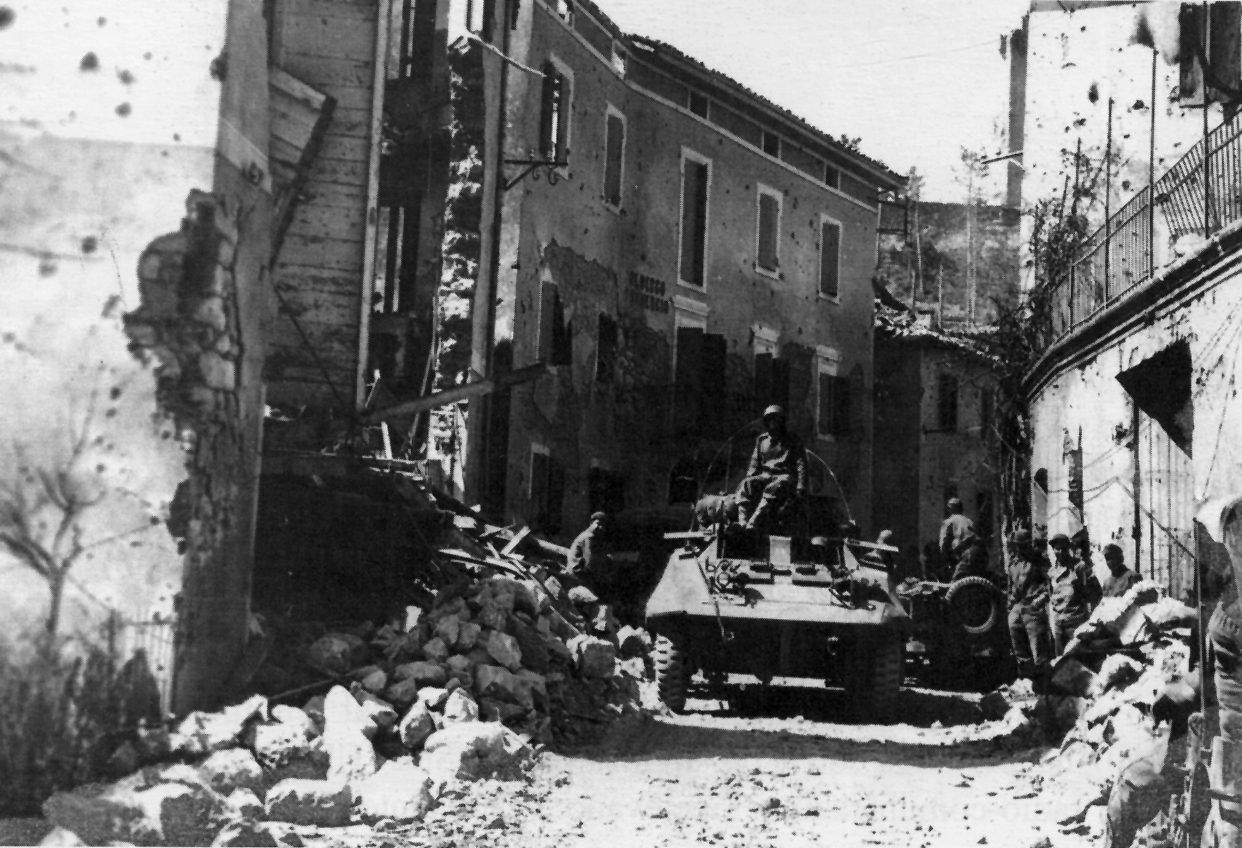 Carro blindado de reconhecimento parado numa rua de Montese, entre casas marcadas por estilhaços e entulho de pedra, com soldados à direita