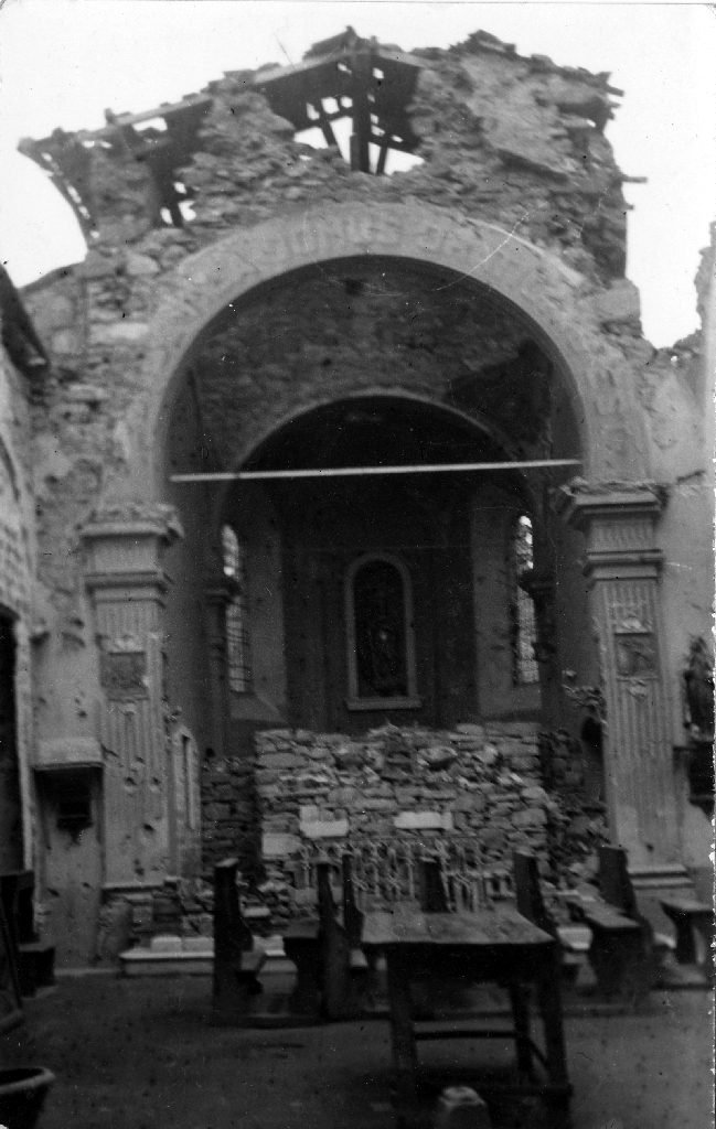 Interior de uma igreja com o teto destruído e a parede do fundo reduzida a entulho, bancos de madeira no chão