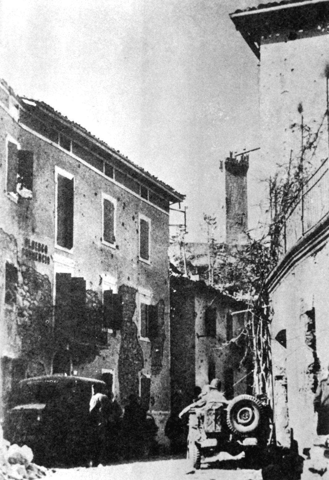 Rua estreita de Montese com fachadas marcadas por estilhaços, um jipe e um caminhão, e a torre da cidade danificada ao fundo