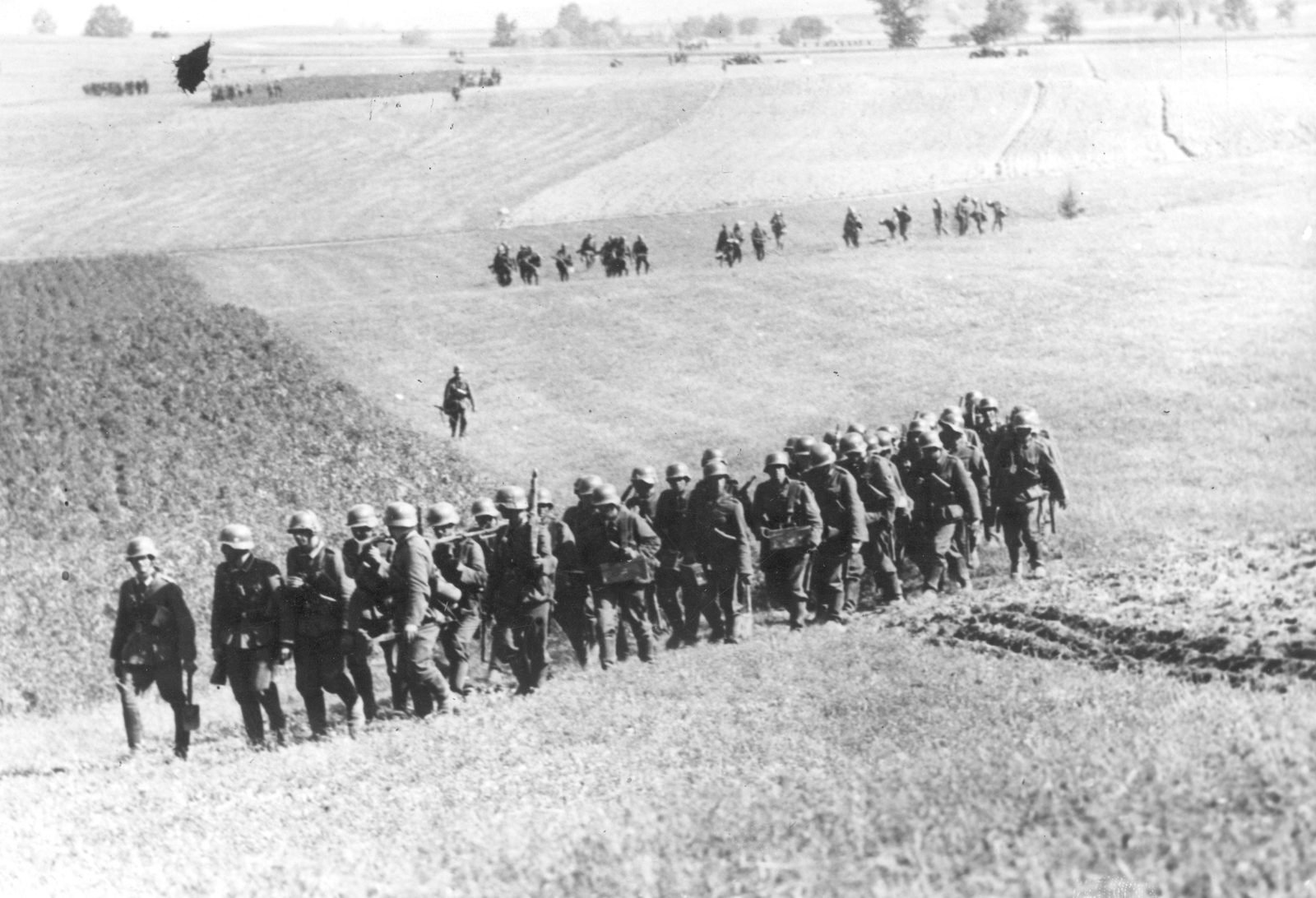 Line of German soldiers in helmets climbing an open hillside on foot, with other groups marching in the background