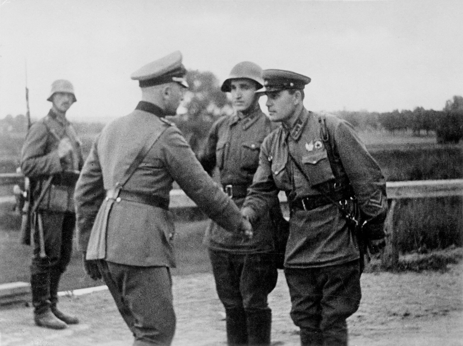 Oficial alemão apertando a mão de um oficial soviético, com outro soldado soviético e uma sentinela alemã ao lado