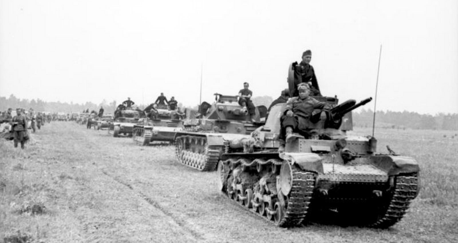 Column of German tanks halted in an open field, with crewmen sitting on the turrets and soldiers on foot alongside