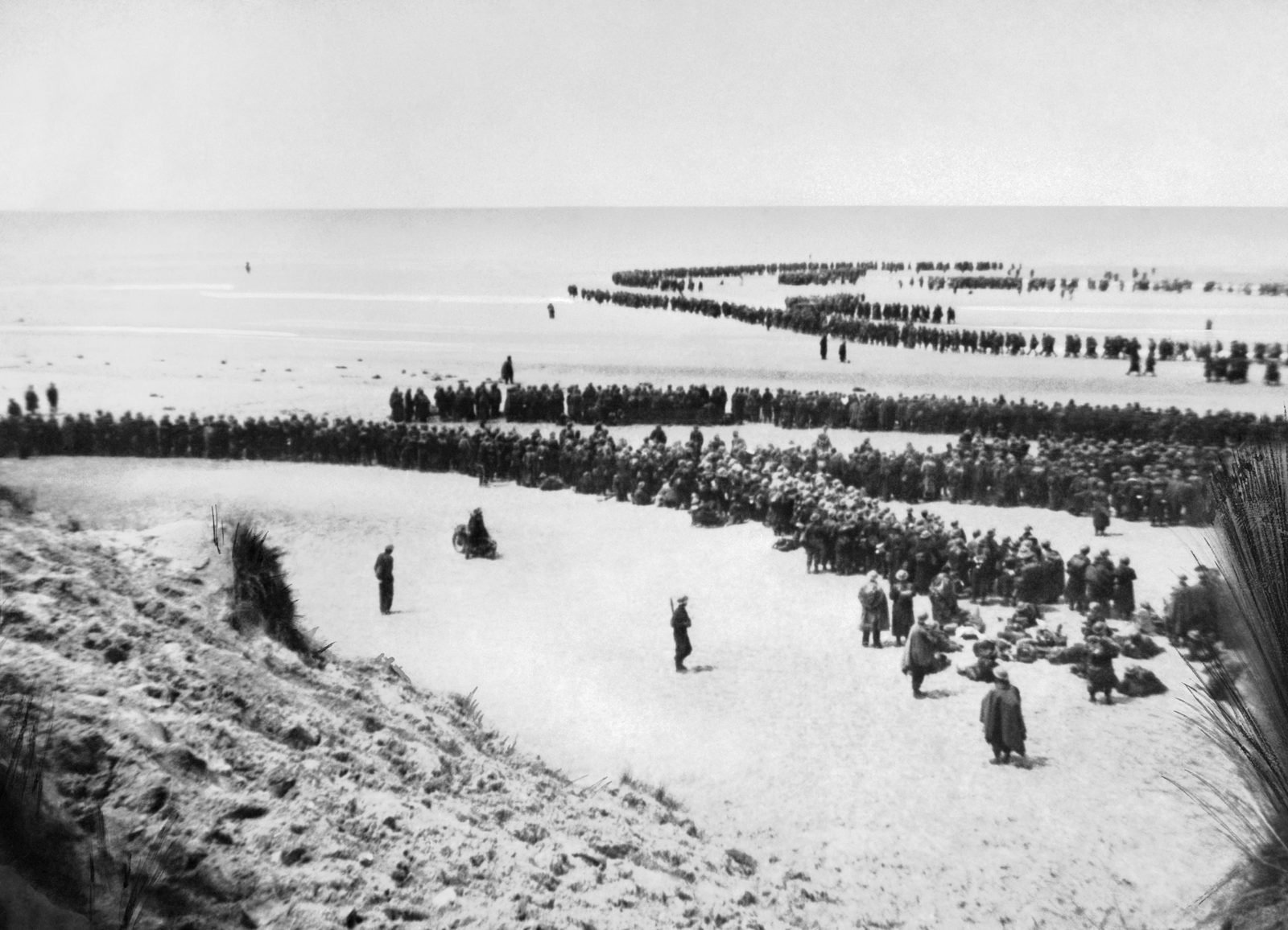 Longas filas de soldados atravessam a praia larga de Dunquerque, das dunas em primeiro plano até a beira da água