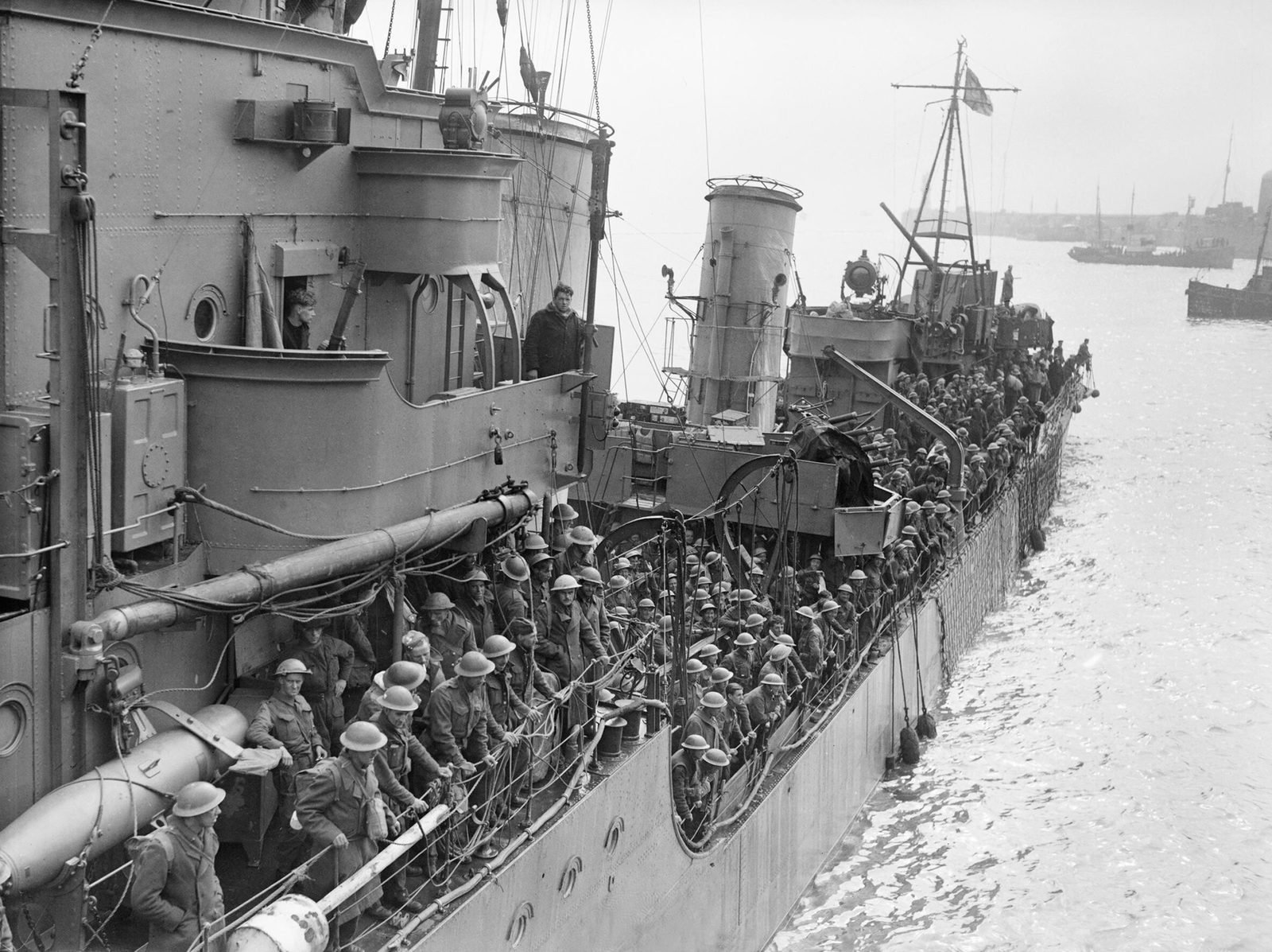 Convés de um contratorpedeiro lotado de soldados britânicos de capacete, encostando num cais, com barcos ao fundo