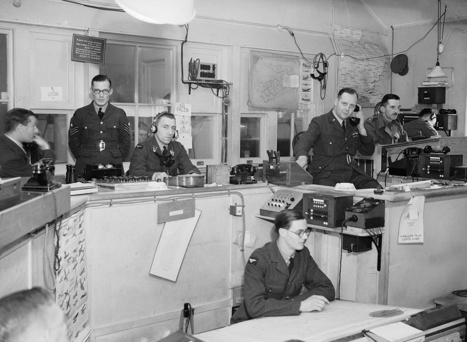 Operations room with men in RAF uniform in front of telephone and radio panels; one officer speaks on the telephone, another wears headphones, and an airman seated in front works at a map table