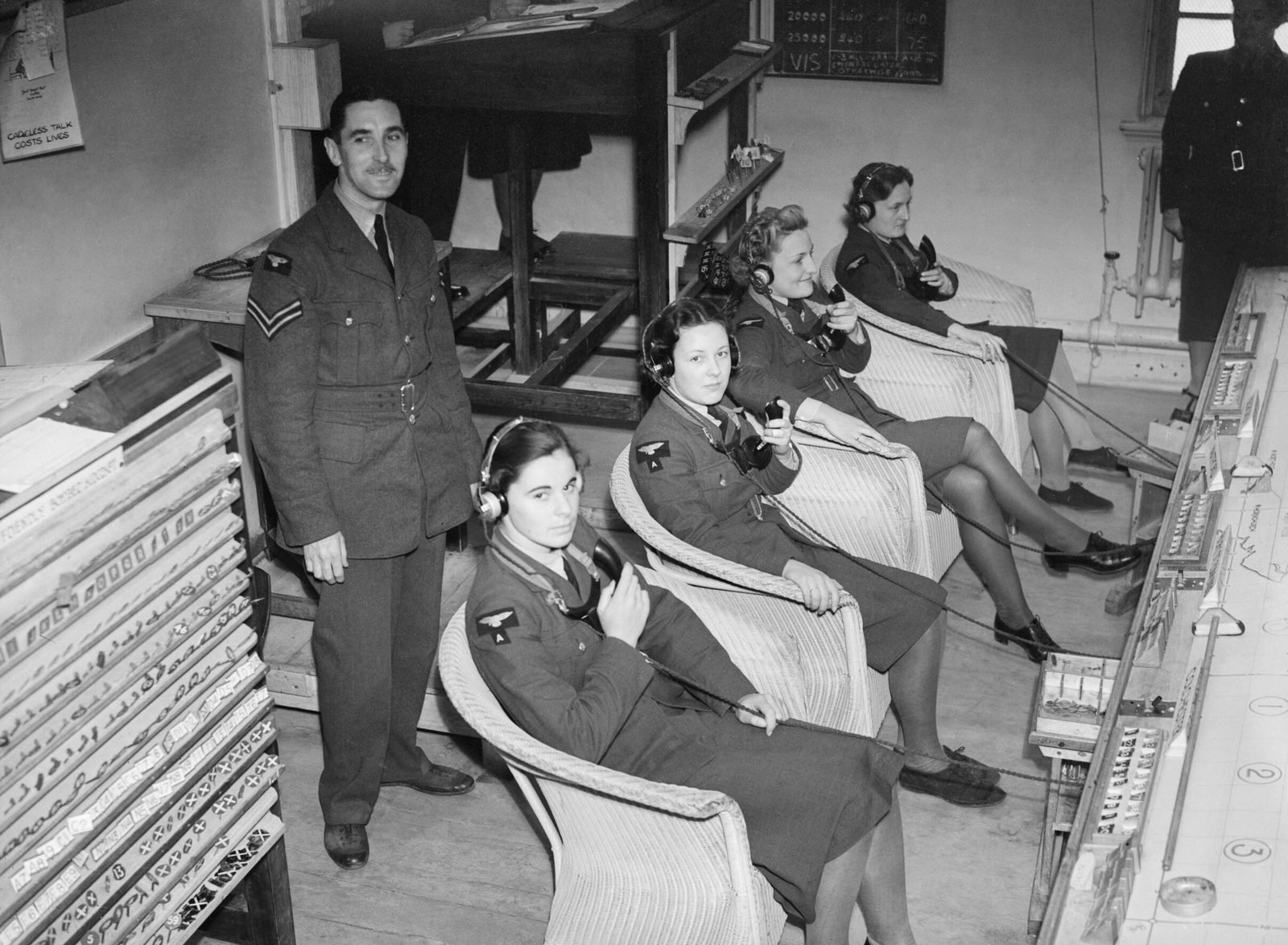 Quatro telefonistas da força aérea feminina, de fones de ouvido, sentadas em poltronas de vime ao lado de uma mesa de plotagem, com um cabo da RAF de pé atrás delas