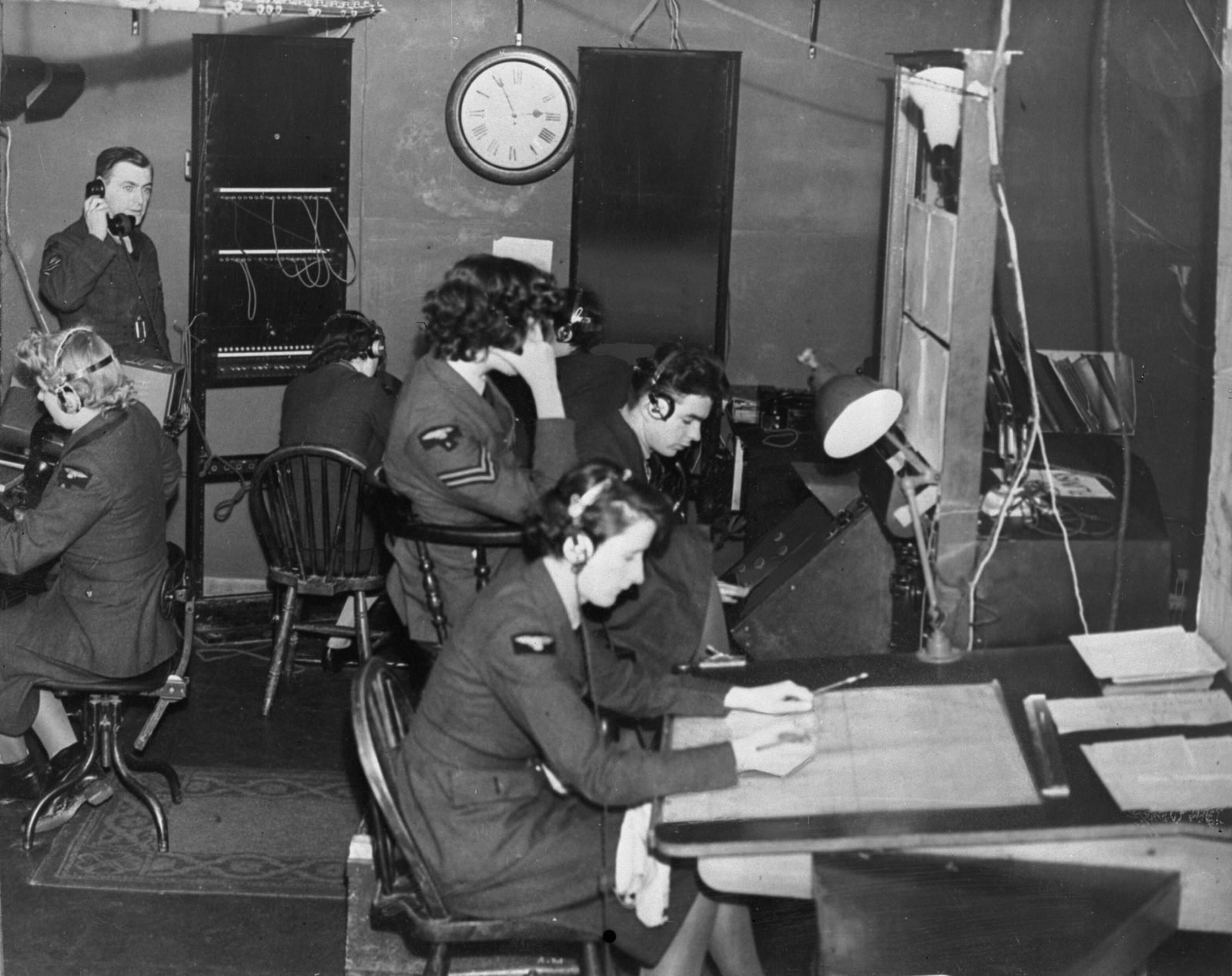 Interior de uma cabana de madeira com operadoras de fones de ouvido sentadas diante de aparelhos e de uma mesa de anotações, um homem ao telefone ao fundo e um relógio na parede