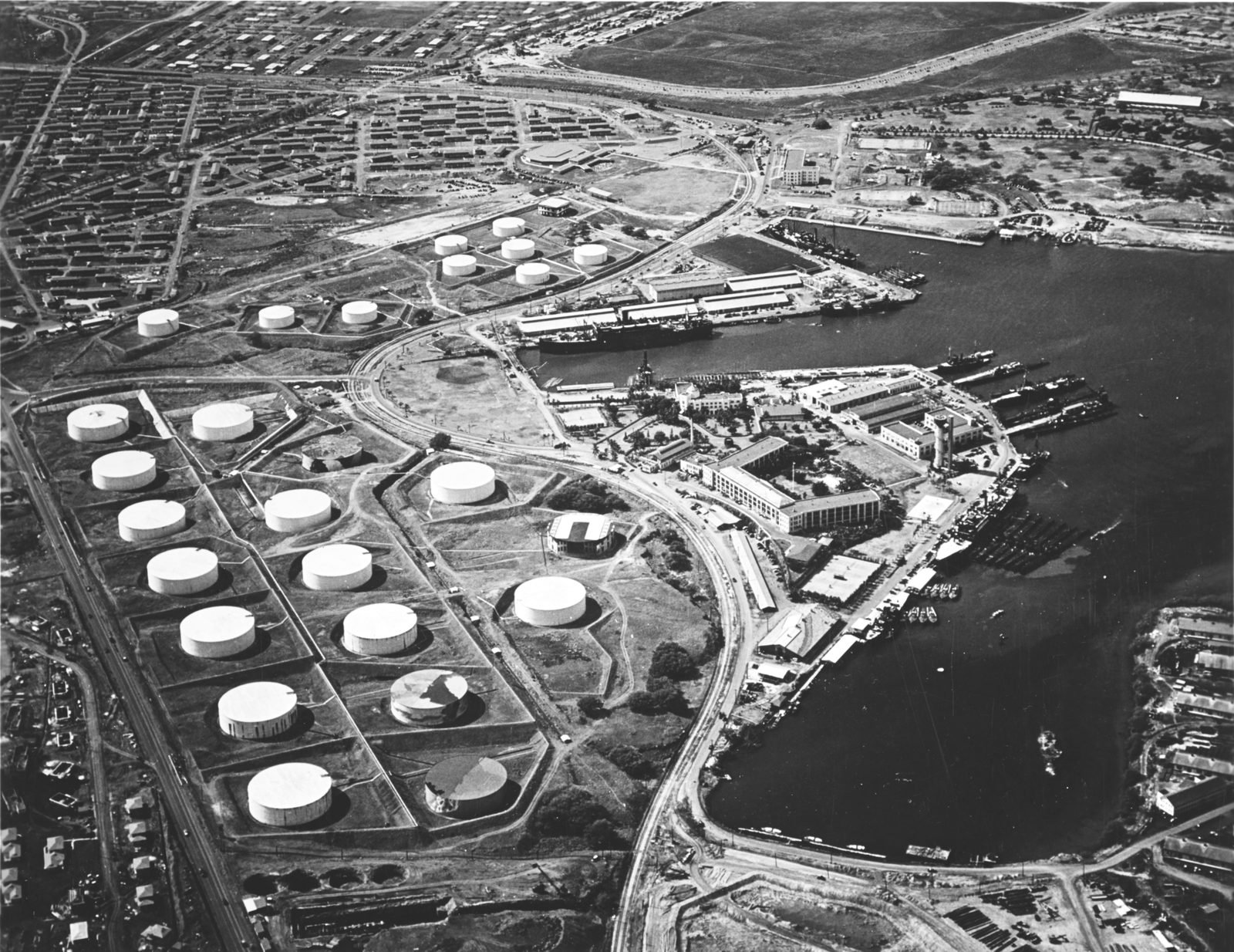 Vista aérea de dezenas de tanques cilíndricos brancos de combustível ao lado de um cais com navios e submarinos atracados