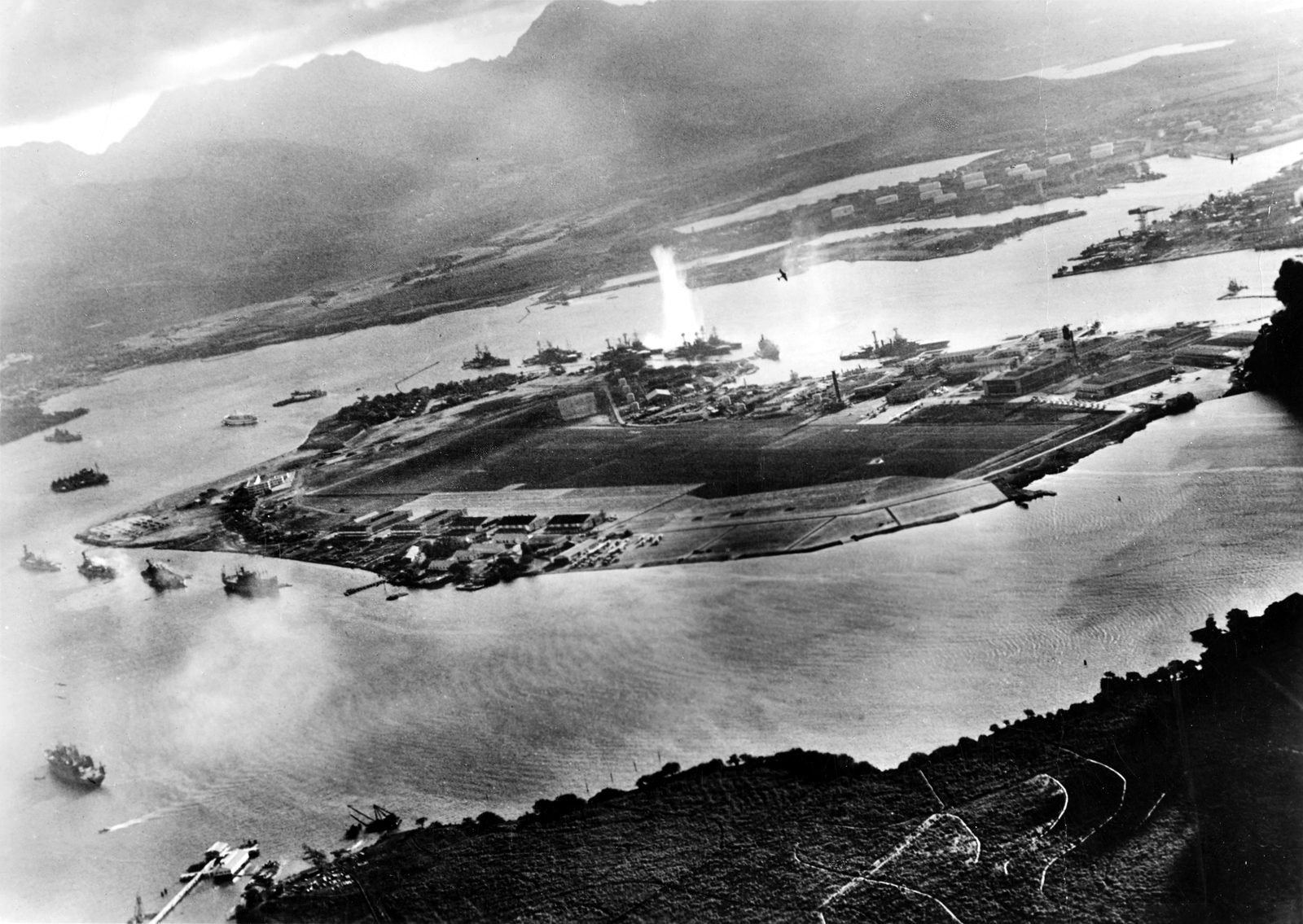 Vista aérea oblíqua de uma ilha plana no meio de um porto, com navios fundeados em fila do outro lado e uma coluna de água subindo junto a um deles