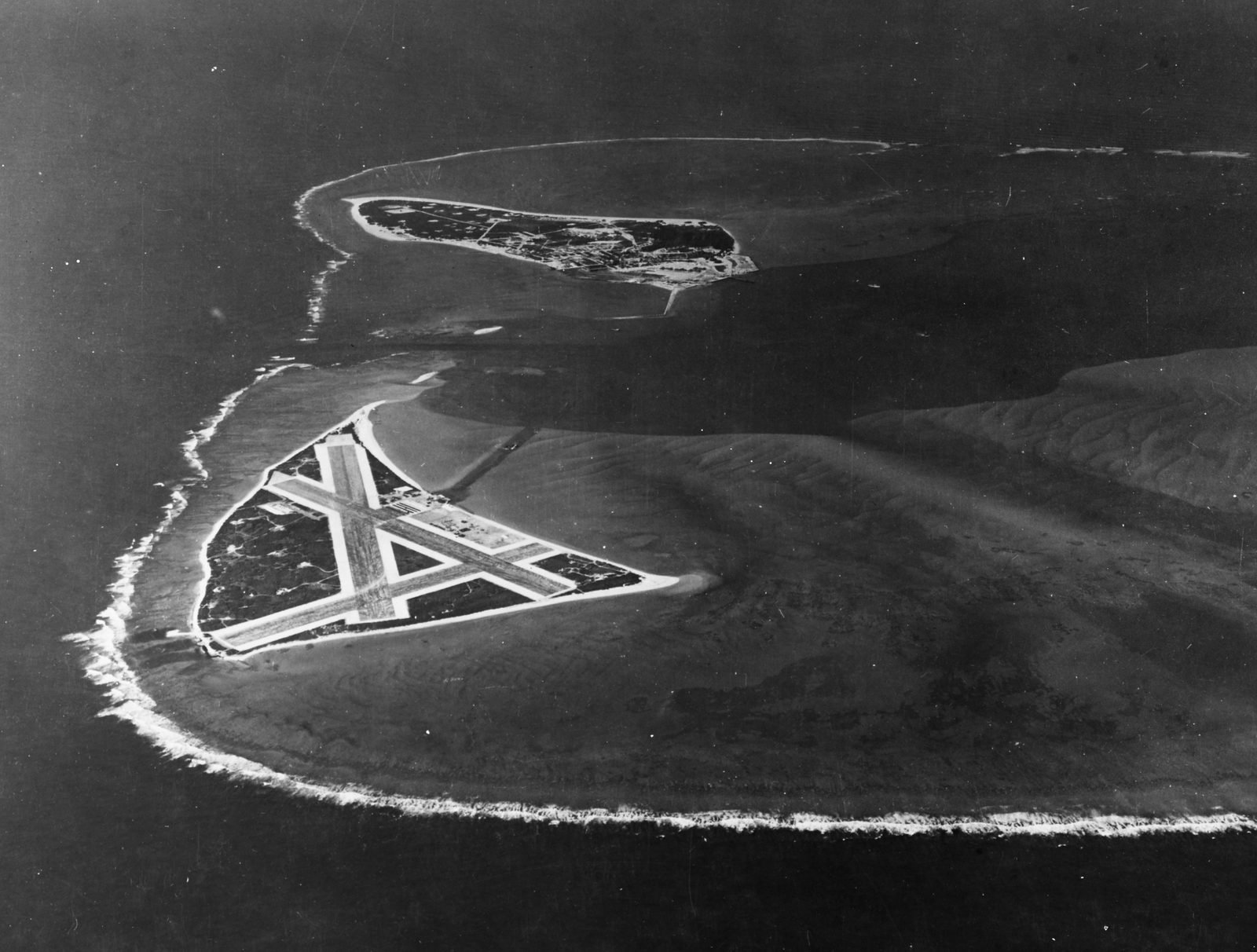 Vista aérea oblíqua de um atol: uma ilha triangular com pistas de pouso em cruz à frente e uma segunda ilha ao fundo, cercadas pela arrebentação do recife