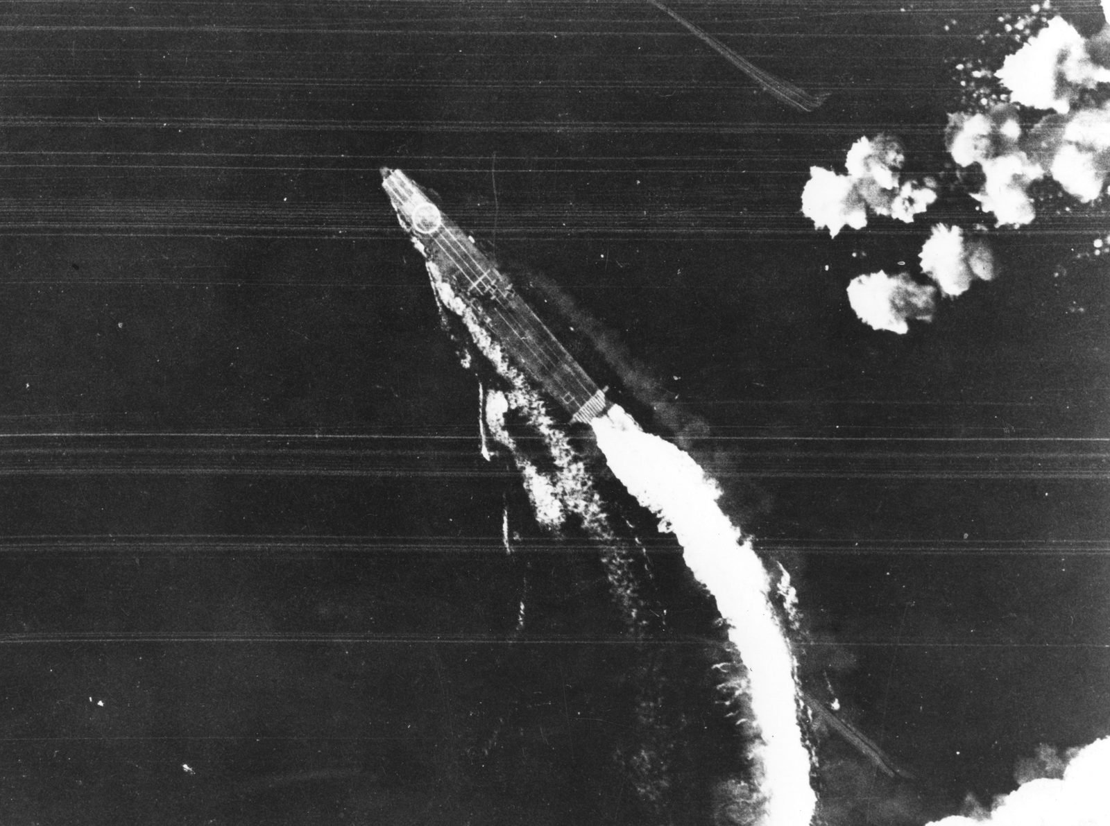 Vista aérea vertical de um porta-aviões fazendo uma curva fechada, deixando uma esteira branca em arco, com explosões de bombas na água à direita