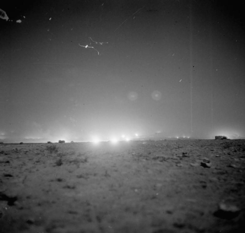 Deserto à noite, com viaturas recortadas contra uma faixa de clarões no horizonte
