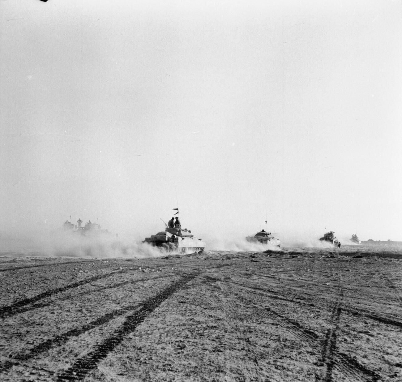 Tanques avançando em fila pelo deserto, levantando poeira, sobre marcas de lagartas no chão
