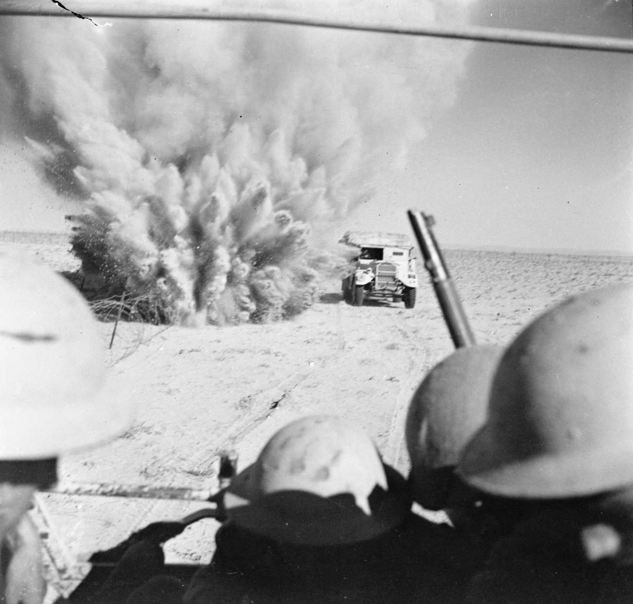 View from the back of a truck with helmeted soldiers seen from behind; ahead, another truck moves through the desert beside a large burst of sand