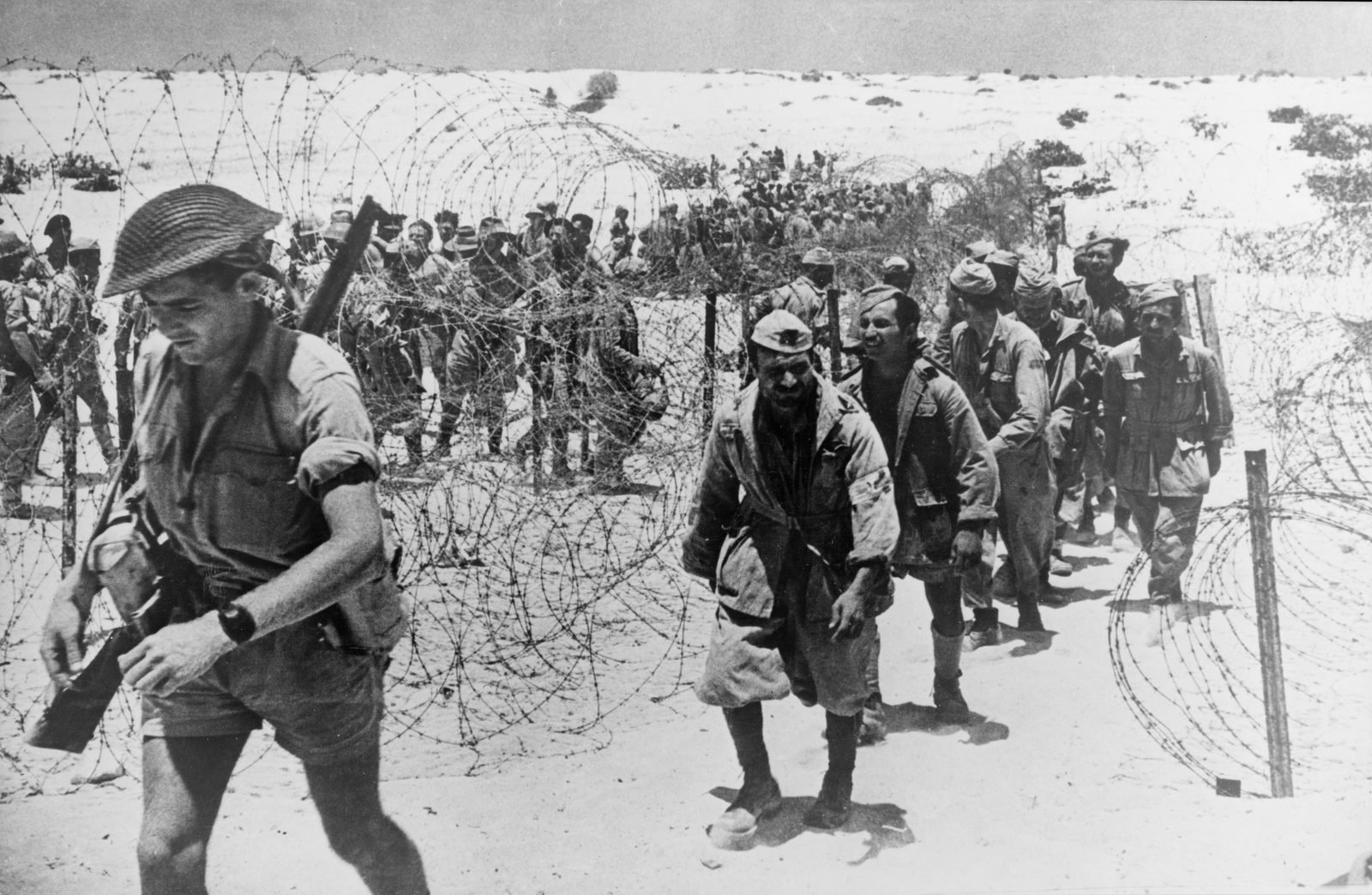 Fila de prisioneiros de guerra caminhando entre cercas de arame farpado, precedida por um soldado britânico de fuzil ao ombro