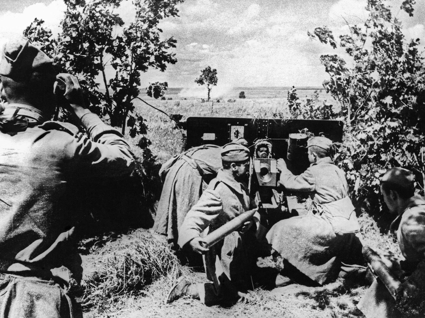 Guarnição soviética de quatro homens em volta de um canhão escondido entre arbustos, um deles segurando uma granada, com a estepe aberta ao fundo