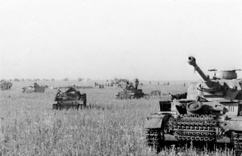 Um tanque alemão Panzer IV em primeiro plano à direita e outros blindados espalhados num campo de trigo sob céu claro
