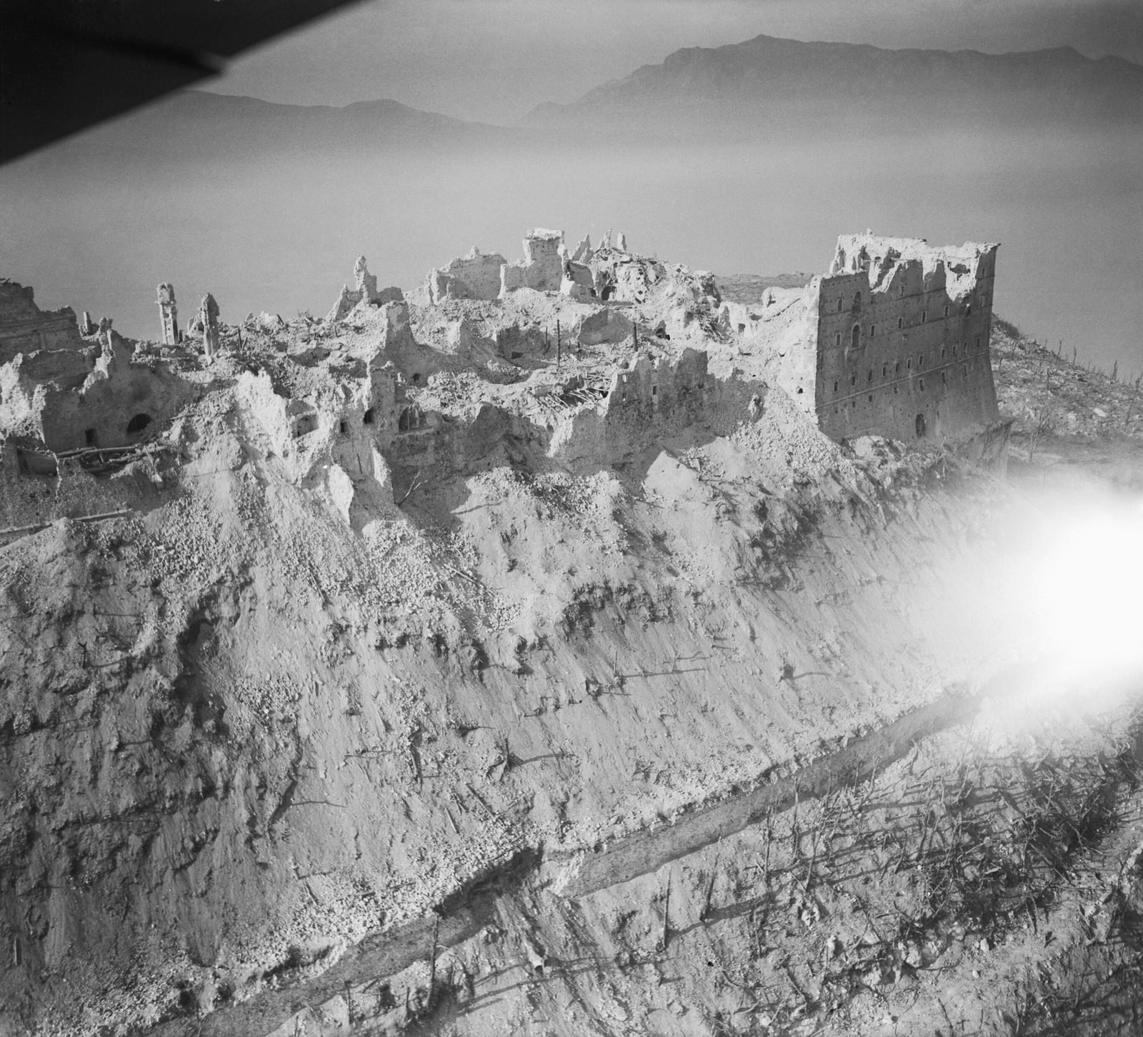 Low aerial view of the ruins of the abbey on top of Monte Cassino: a stone wall still standing on the right, rubble over the rest of the summit and mountains in the background