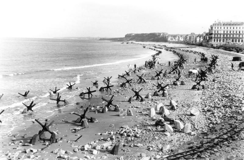 Praia longa com fileiras de obstáculos antitanque de vigas de aço cruzadas e blocos de concreto, com prédios à beira-mar e uma falésia ao fundo