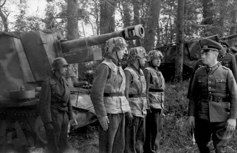 Um marechal de quepe e uniforme fala com soldados de capacete camuflado, enfileirados diante de um canhão autopropulsado num bosque