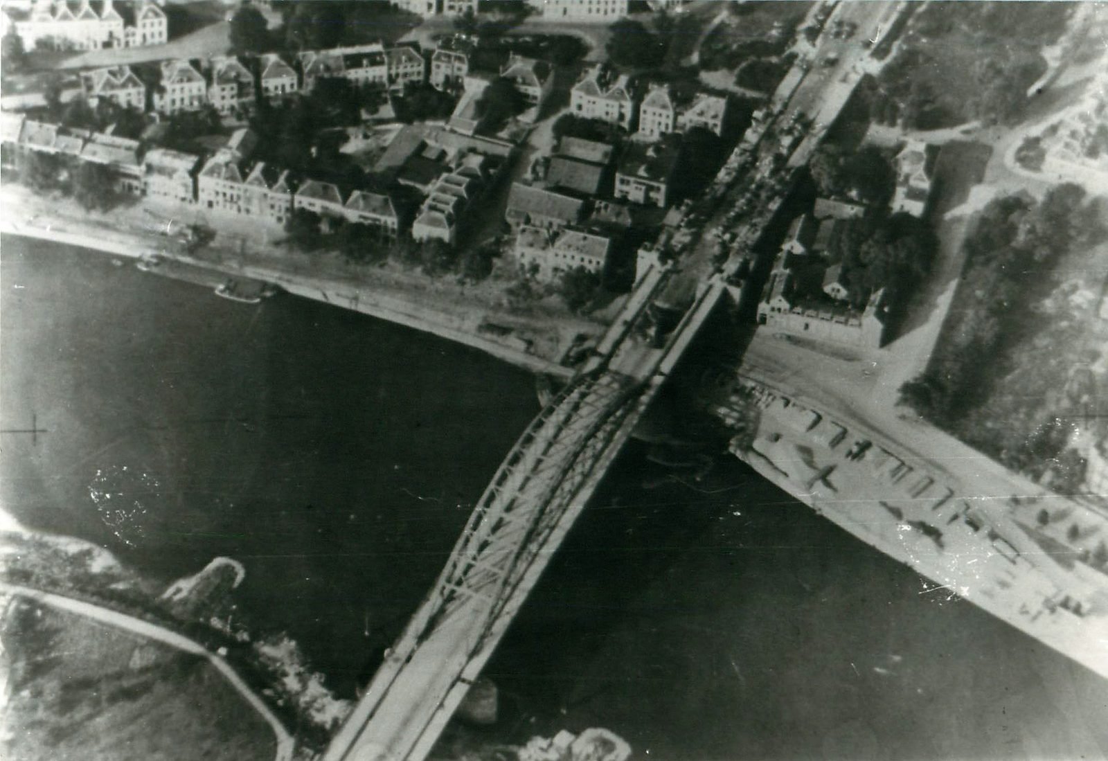 Aerial view of a steel arch bridge over a wide river, with houses on the upper bank and destroyed vehicles piled on the ramp at the northern end