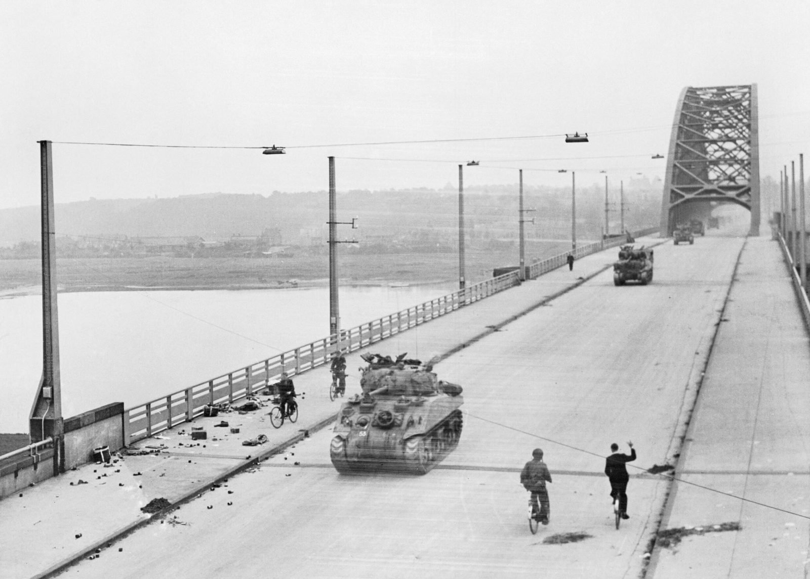 Tanque Sherman avança por uma ponte rodoviária larga, com um grande arco de aço ao fundo e dois ciclistas em primeiro plano, um deles acenando