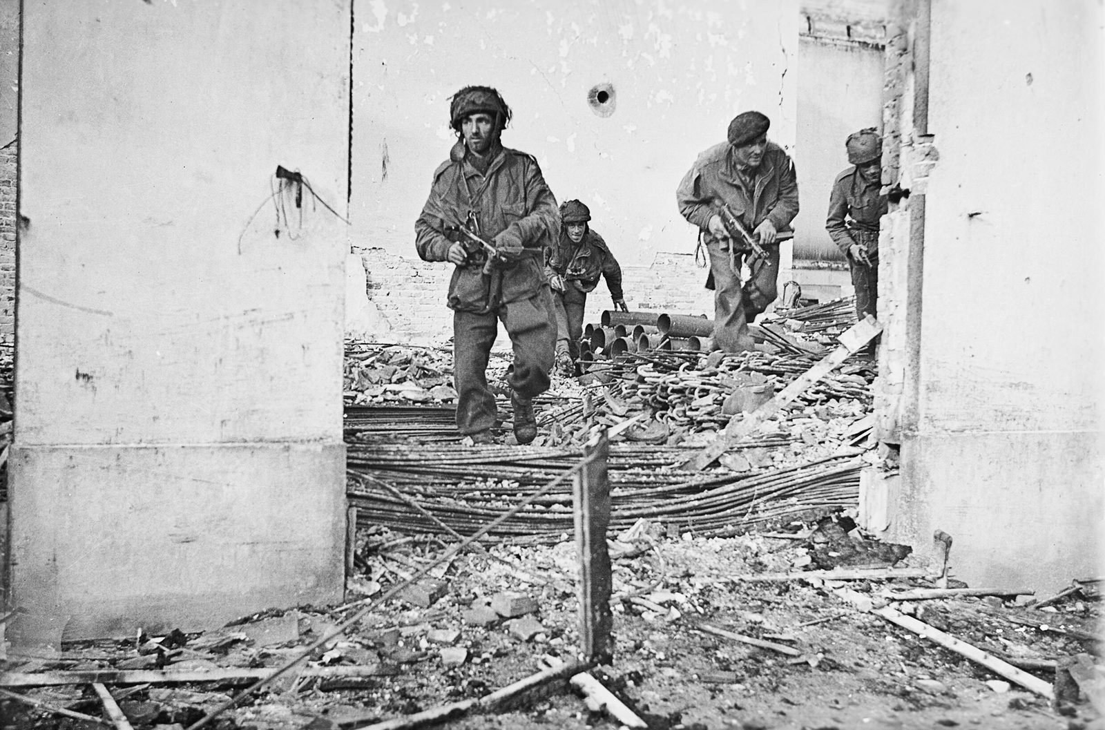 Quatro paraquedistas britânicos armados avançam por uma casa em ruínas, pisando em entulho e vergalhões