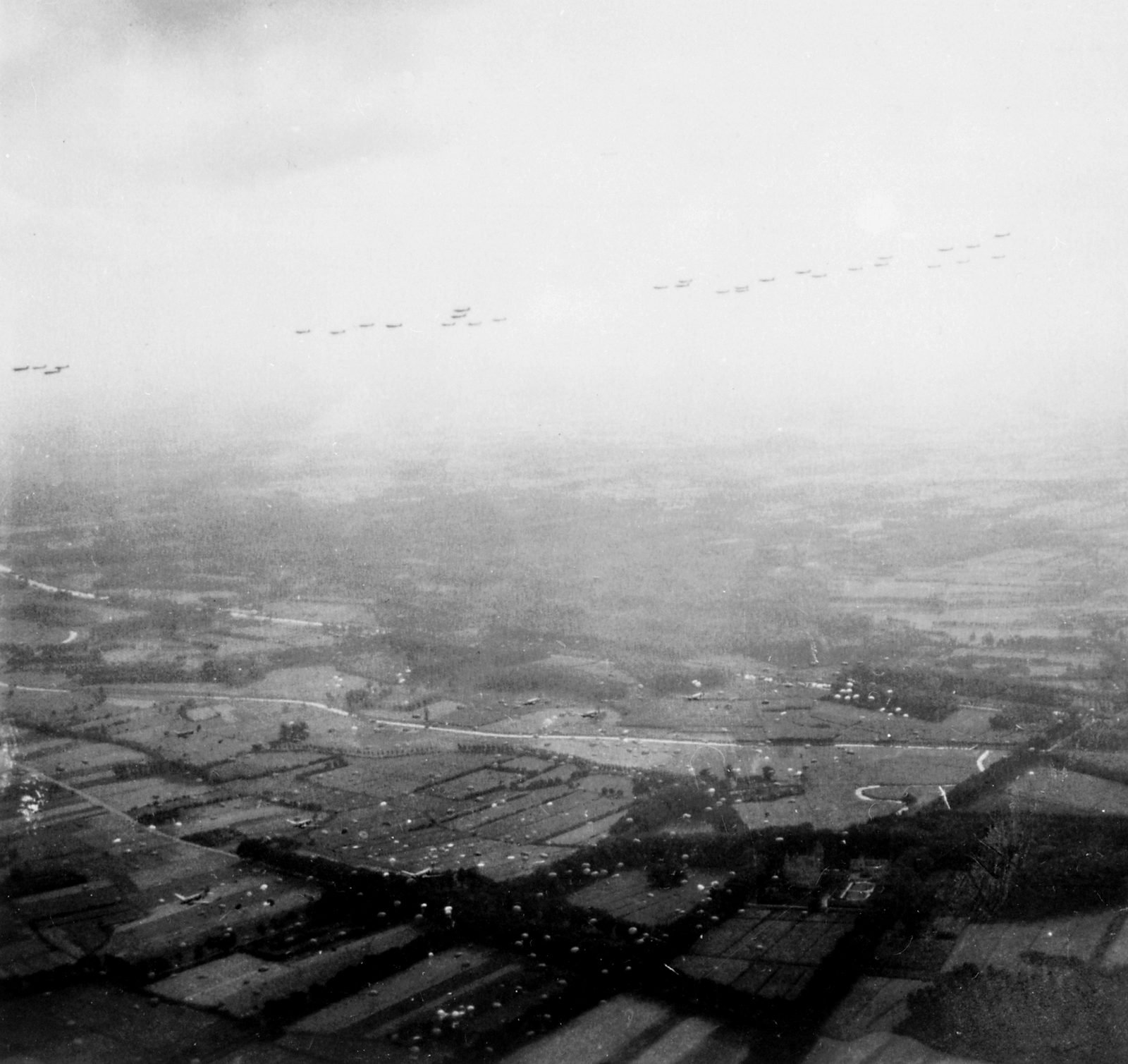 Fotografia aérea em preto e branco: uma longa fileira de aviões de transporte no céu enevoado e, embaixo, campos retalhados salpicados de paraquedas abertos
