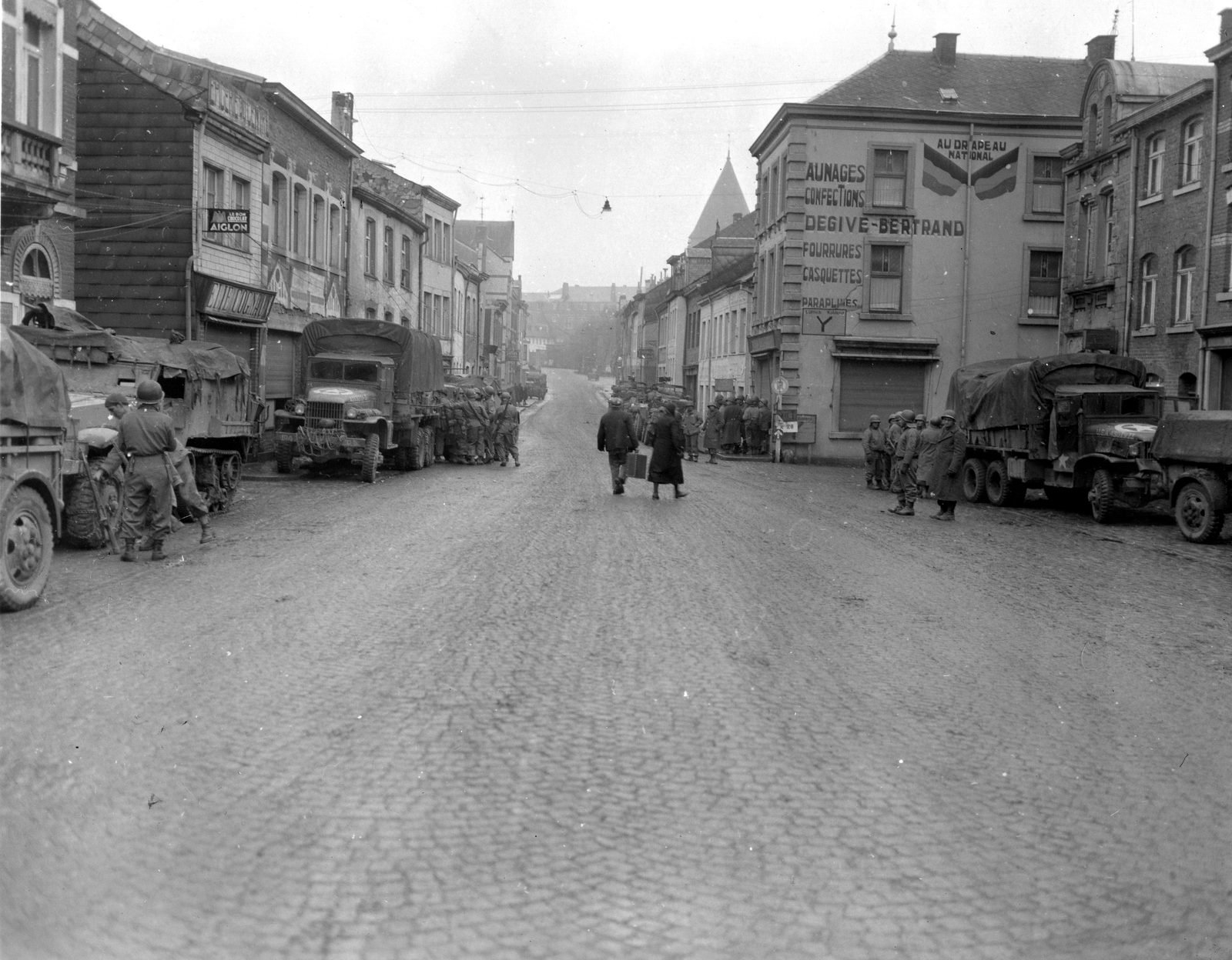 Rua de paralelepípedos em Bastogne com caminhões militares estacionados dos dois lados, soldados agrupados e um casal de civis carregando malas no meio da rua