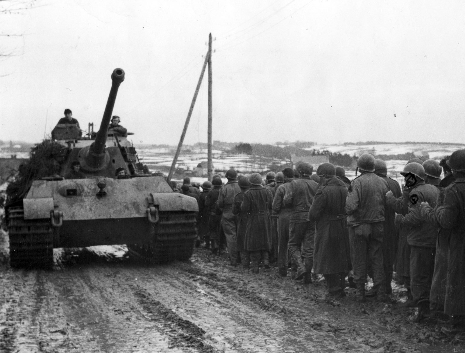 Um tanque pesado alemão avança por uma estrada enlameada com neve nos campos, ao lado de uma longa fila de soldados americanos de capacete, prisioneiros