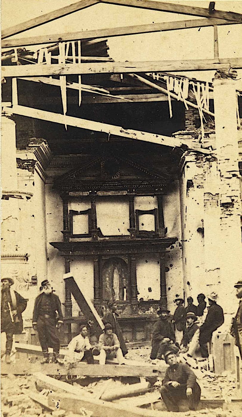 Interior de uma igreja destruída, com o telhado aberto em vigas quebradas, o retábulo do altar ao fundo e soldados de chapéu sentados e de pé sobre os escombros