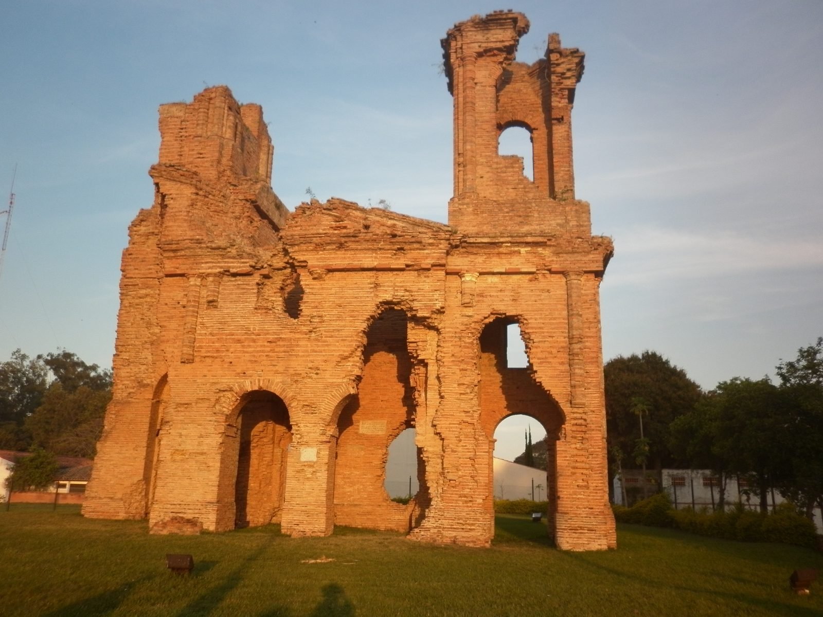 Ruínas de tijolo de uma igreja, com arcos abertos e uma torre partida, sobre um gramado à luz do fim de tarde